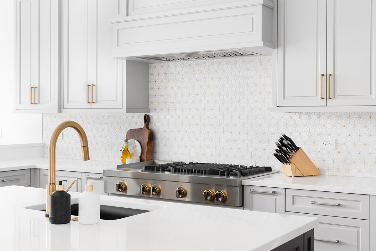 A beautiful kitchen's stainless steel luxury stove, hood, granite counter tops, and a custom, white and gold tiled back splash and faucet.